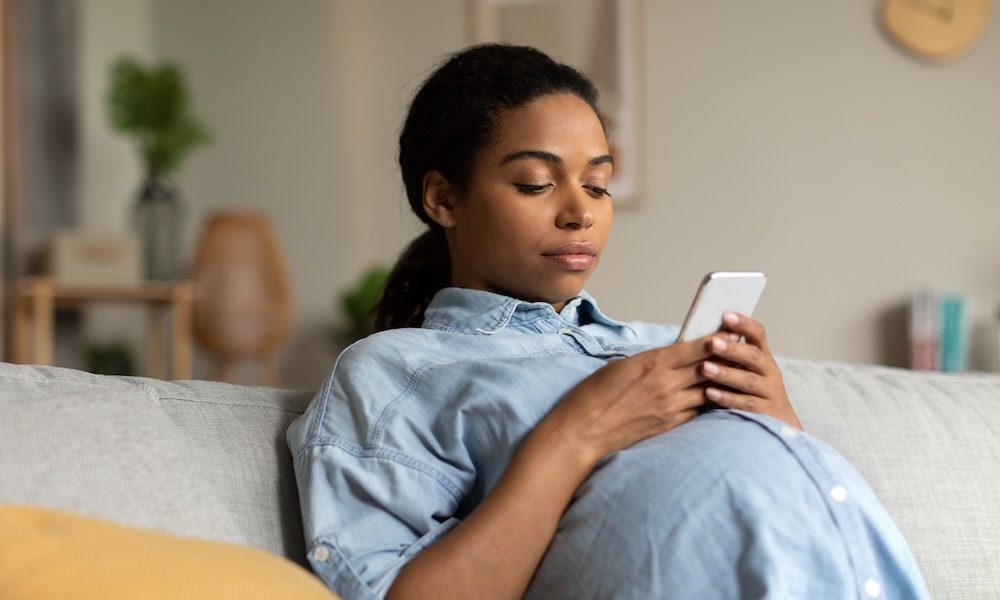 Pregnant Black Lady Using Cellphone Sitting On Sofa At Home AI solution supports maternal and postpartum wellness journey